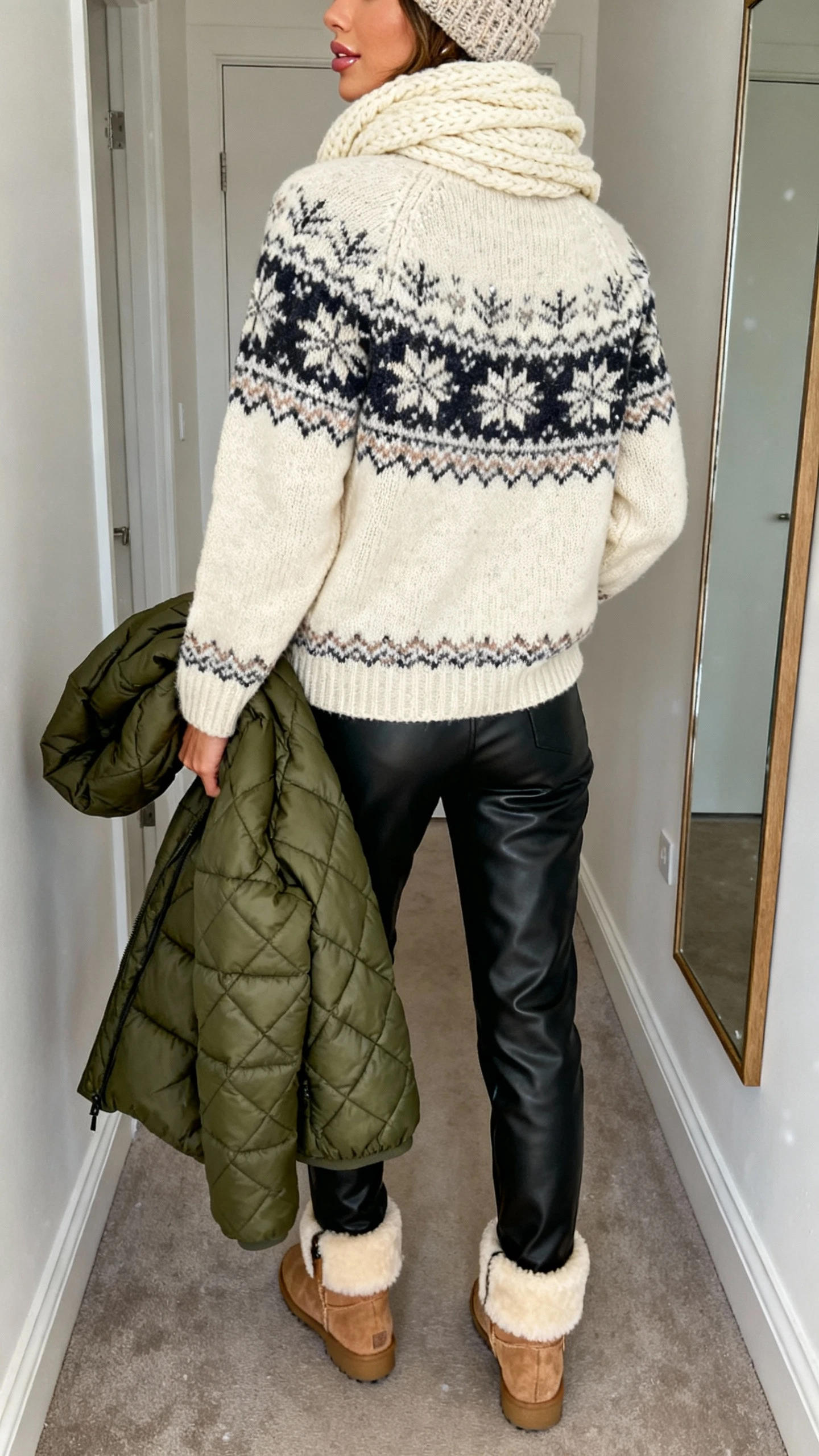 iPhone back view of a woman wearing a classic Fair Isle knit sweater in cream with wintery pattern, black leather pants, chunky cream scarf, shearling-lined ankle boots, quilted puffer coat in forest green over one arm, knit beanie, subtle glowy makeup and glossy lip for snow-kissed glam, face not visible, hallway mirror, natural daylight, iPhone photo quality.